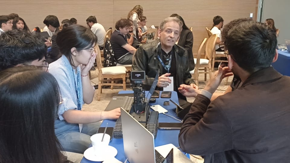 Marcos Moreira Alves seated at a table advising a student team, with laptops and a small robot on the table.