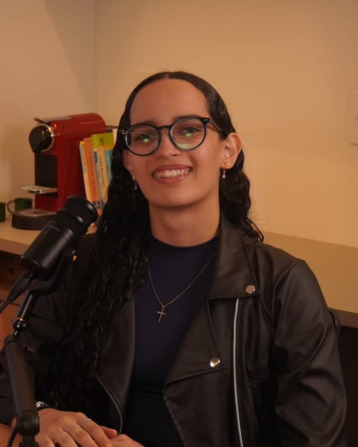 Sophia Oliveira Alves smiling, wearing glasses and a black jacket, seated in front of a studio microphone.